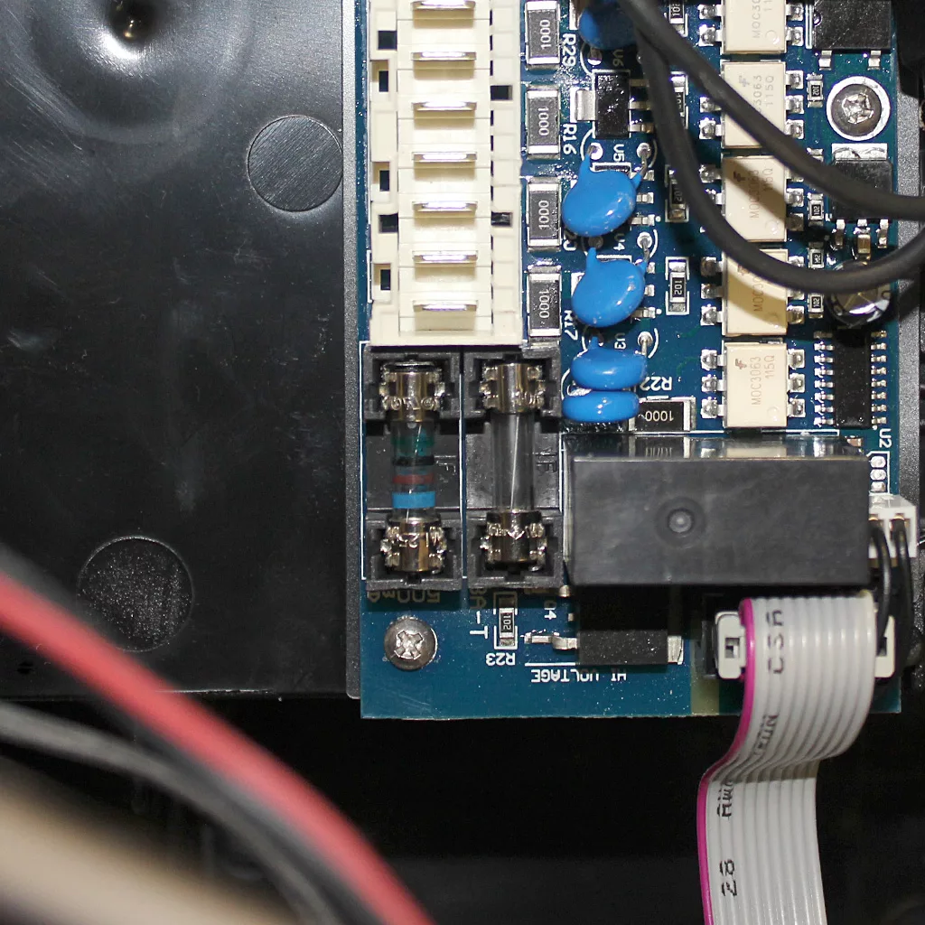 Close-up of an electronic circuit board for the Faema Carisma, featuring blue capacitors, a fuse upgrade, a row of white connectors, relay, fuses, and a ribbon cable in the lower right corner. © 2025 1st-line.com