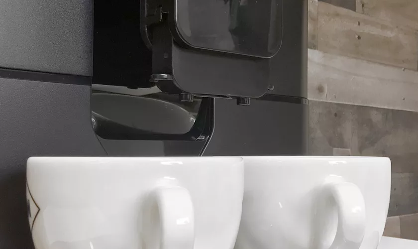 Two white ceramic cups sit under the spouts of a DISCONTINUED Saeco New Royal OTC Super-Automatic Espresso Machine, set against a warm, wood-textured wall, ready to be filled. © 2025 1st-line.com