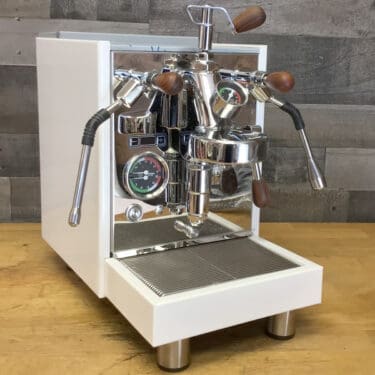 A RETURNED & REFURBISHED Bezzera Aria HX Special Edition Espresso Machine with flow control and chrome accents sits on a wooden countertop against a wood-paneled wall. © 2025 1st-line.com