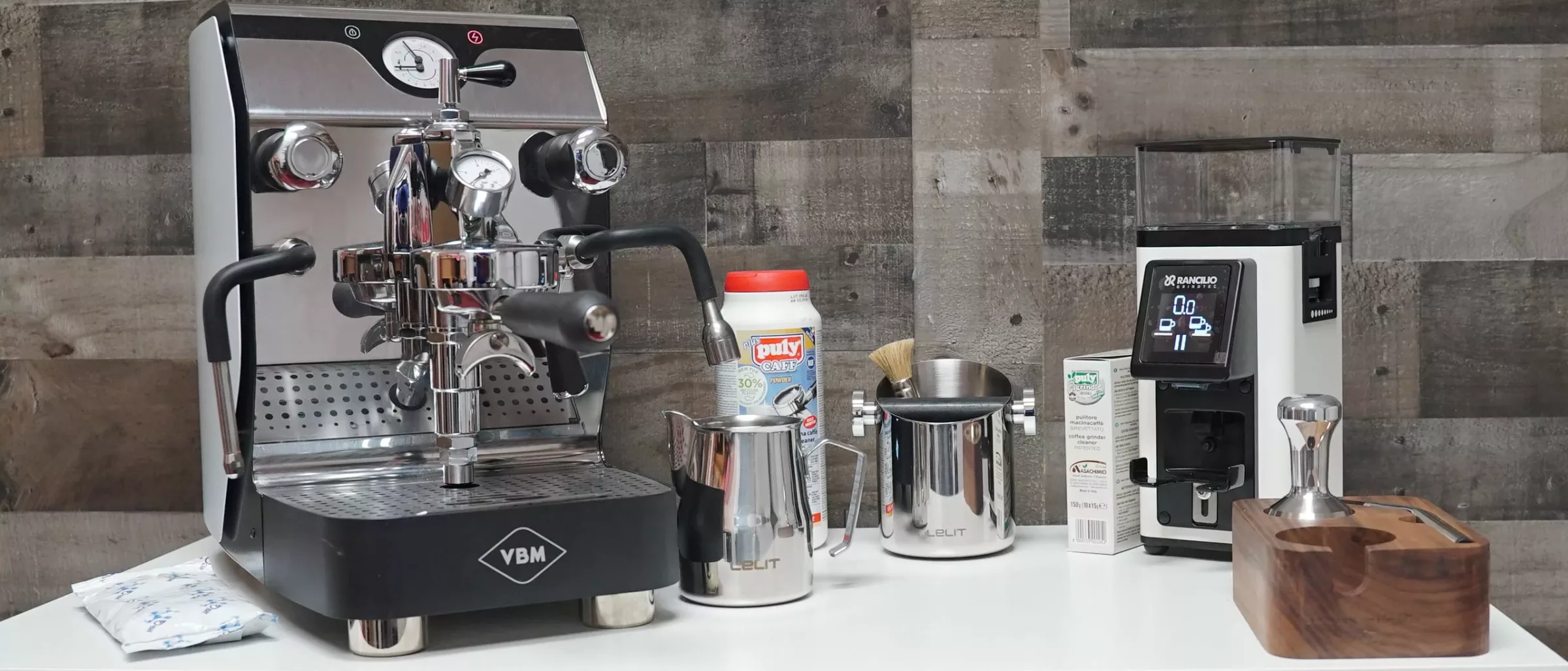 An espresso machine and a coffee grinder sit on a counter with a SupManAna milk frother jug, coffee tamper, milk carton, powdered creamer, and a small brush, all against a wooden wall backdrop. © 2025 1st-line.com