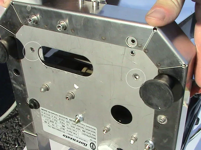 Close-up view of the underside of a Fiorenzato Bricoletta appliance, showing screws, bolts, a label, and two black rubber feet. A person’s hands are holding the edges, possibly preparing for OPV adjustment. © 2025 1st-line.com