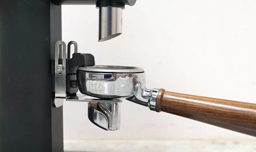 A close-up of a portafilter with a wooden handle on an espresso machine sits beside the Pinecone Pinion All-Purpose Espresso Coffee Grinder, both ready for brewing coffee. © 2025 1st-line.com