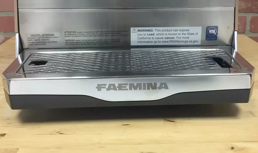 A close-up of the Faema Faemina Dual Boiler Espresso Machine tray, featuring a chemical exposure warning label, placed on wood with a brick wall backdrop. © 2025 1st-line.com