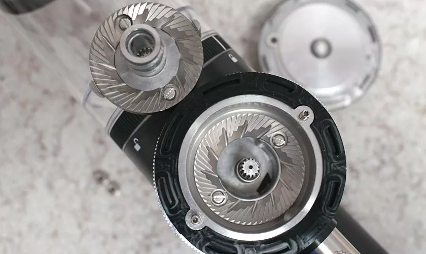 Close-up of a disassembled Pinecone Pinion All-Purpose Espresso Coffee Grinder on a light speckled surface, displaying exposed burrs with intricate metal teeth and both parts of the burr mechanism. © 2025 1st-line.com