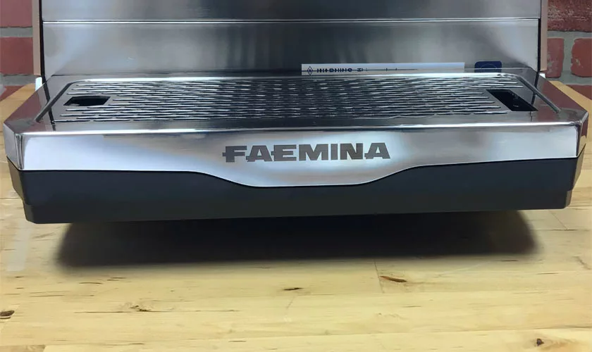 Close-up of the stainless steel drip tray and grate of the Faema Faemina Dual Boiler Espresso Machine on a wooden countertop, backed by a brick wall. This machine blends modern design with timeless charm. © 2025 1st-line.com