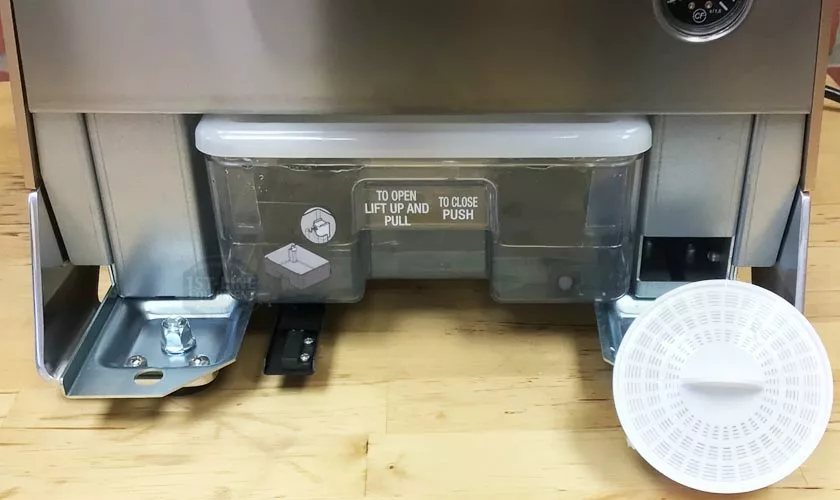 Close-up of the transparent water reservoir attached to the Faema Faemina Dual Boiler Espresso Machine, showing instructions for opening and closing. A round white grated plastic component rests on the wooden surface nearby. © 2025 1st-line.com