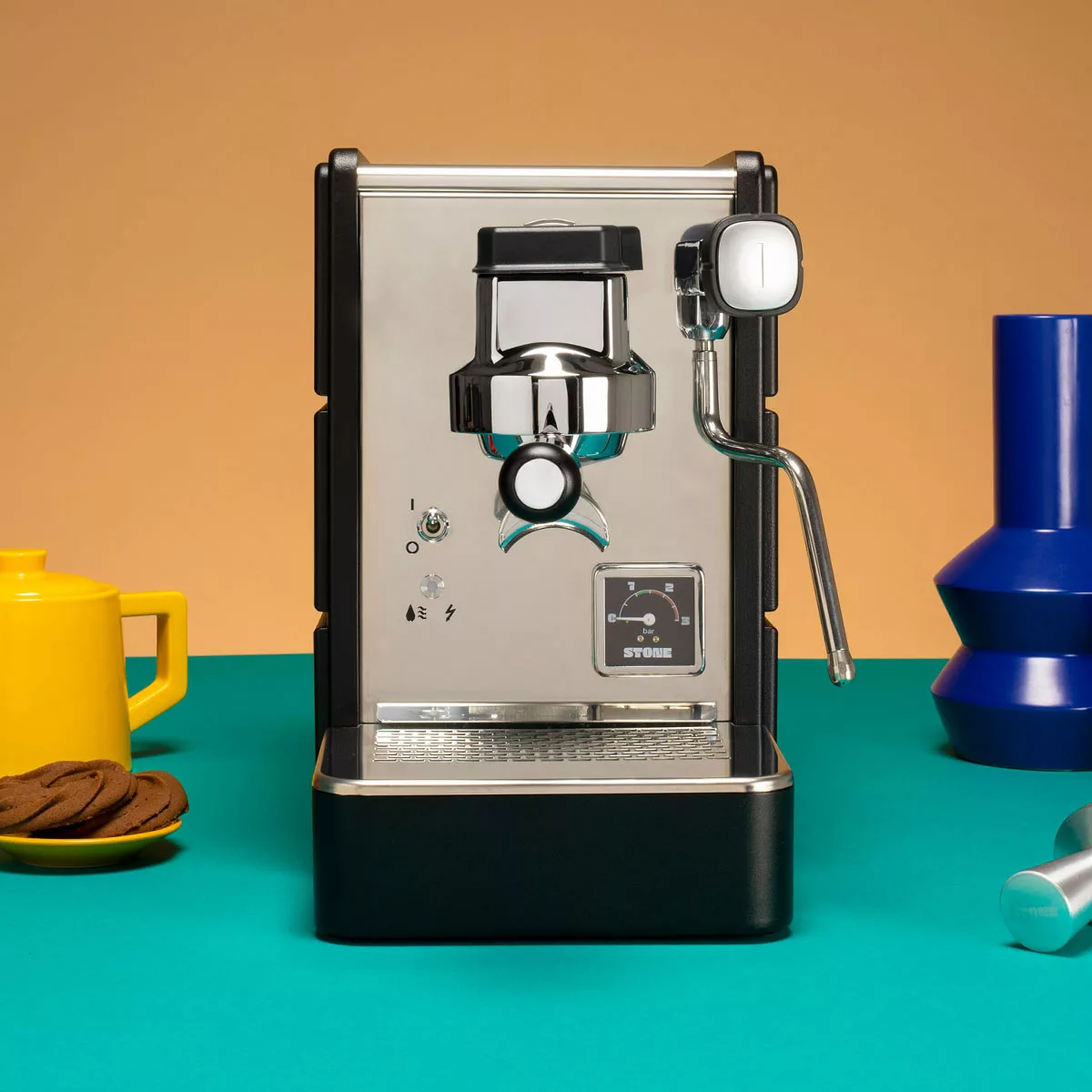 The Stone HX Espresso Machine, featuring a metallic front and black sides, sits on a teal surface with a yellow mug, cookies, and a blue vase nearby, all set against a peach-colored background. © 2025 1st-line.com