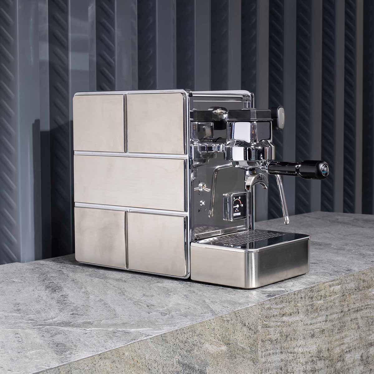 The Stone HX Espresso Machine, featuring a stainless steel finish, steam wand, and portafilter for professional-quality brewing, sits on a stone countertop against gray textured panels. © 2025 1st-line.com