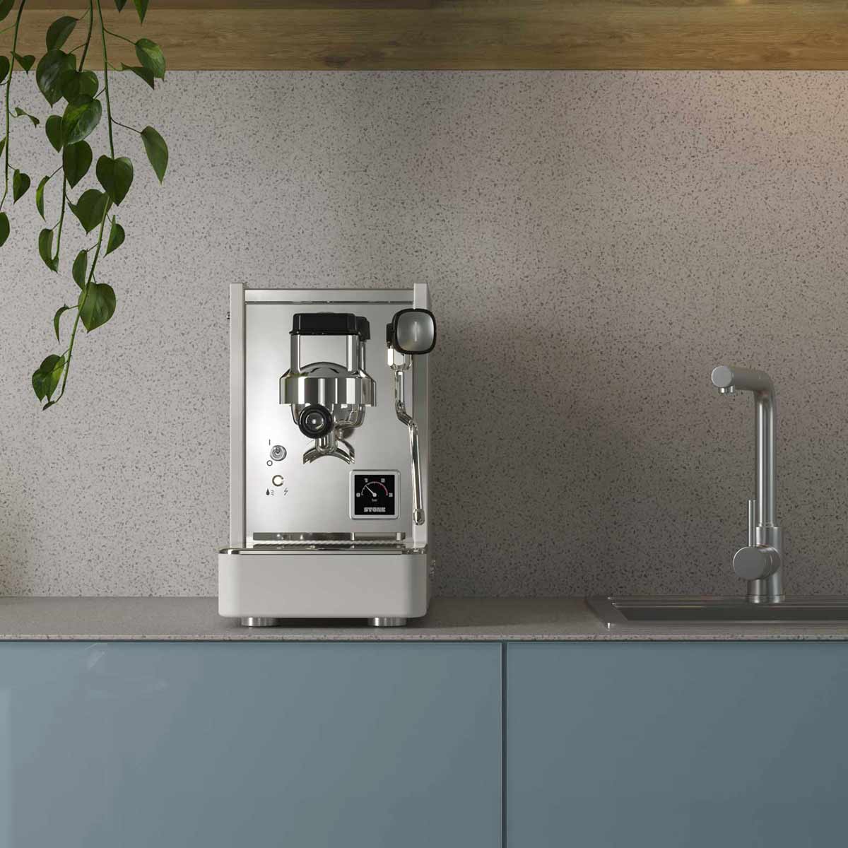 The Stone HX Espresso Machine in silver stands on a grey countertop beside a modern sink, while green hanging plant leaves descend from above against a speckled, light-colored wall. © 2025 1st-line.com
