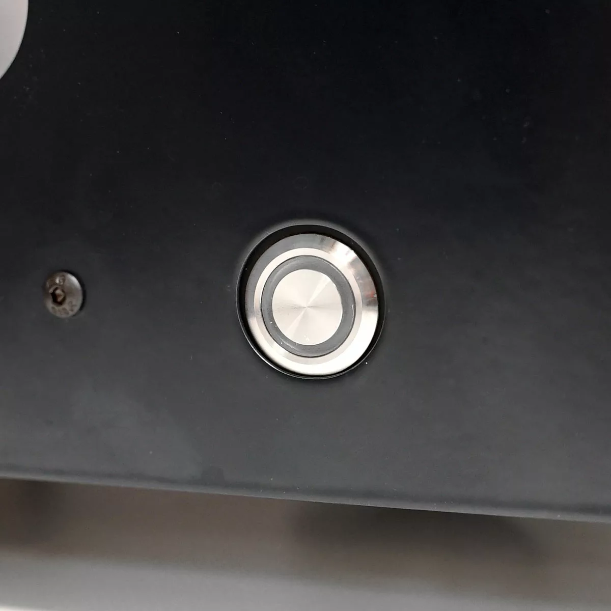 A close-up of a round metallic push button set into the dark panel of a VBM Domobar Super, with a small screw visible to the left of the button. © 2025 1st-line.com