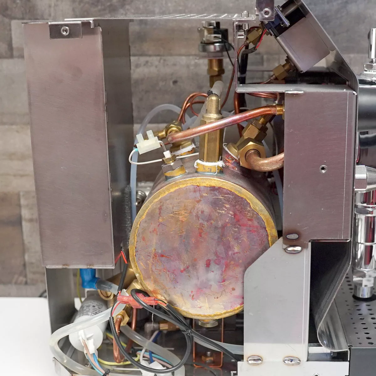 A close-up view of the internal components of a VBM Domobar Super coffee machine, showing metal casing, wiring, pipes, and a cylindrical brass boiler, with the side panel removed for visibility. © 2025 1st-line.com