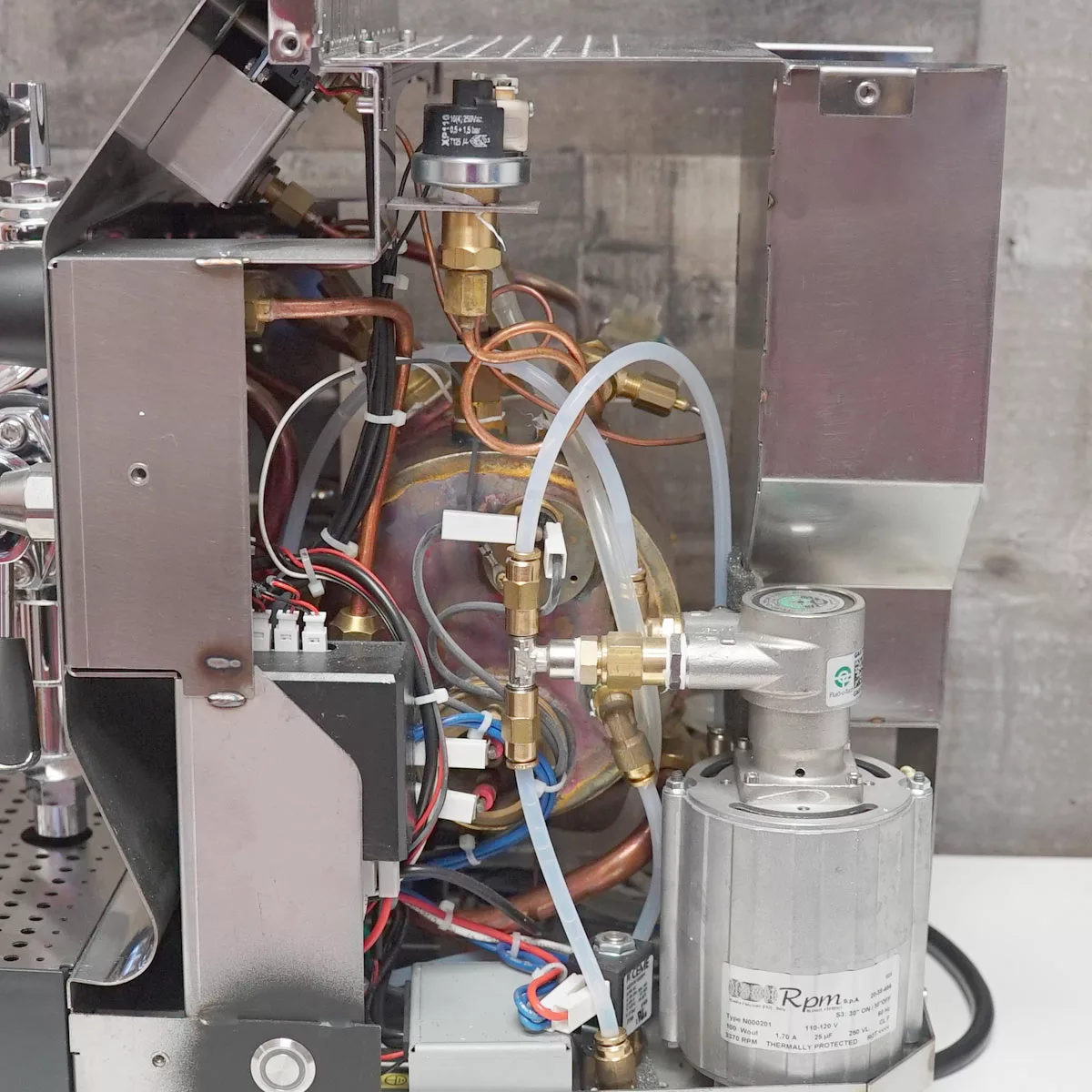 A partially open VBM Domobar Super espresso machine reveals its internal components, including wires, tubes, valves, and a metal pump, with a gray panel on the right and various metal and plastic parts visible for inside access. © 2025 1st-line.com