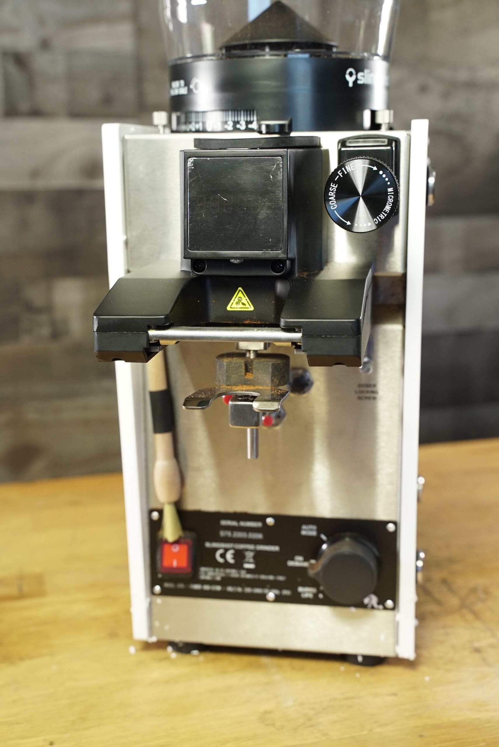 A close-up of the CONVENTION DEMO - Slingshot Dosis S75 Espresso Coffee Grinder White 220V featuring a digital display, control knobs, and coffee chute on a wooden surface with a blurred background; made for 220V commercial use. © 2025 1st-line.com