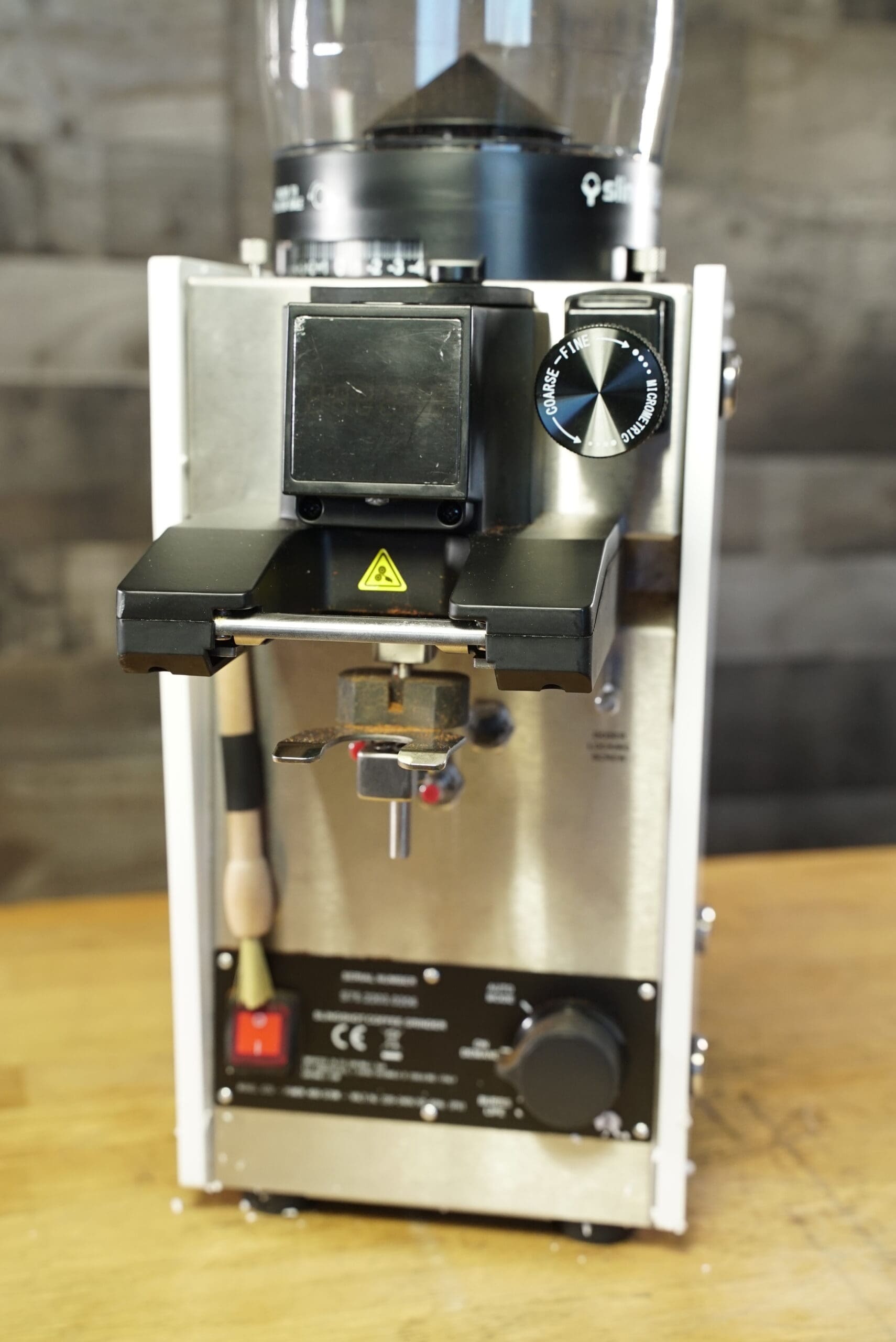 A close-up of the CONVENTION DEMO Slingshot Dosis S75 Espresso Coffee Grinder White 220V, featuring a metal body, control knobs, digital display, and hopper on top, placed on a wooden surface with a blurred background. © 2025 1st-line.com