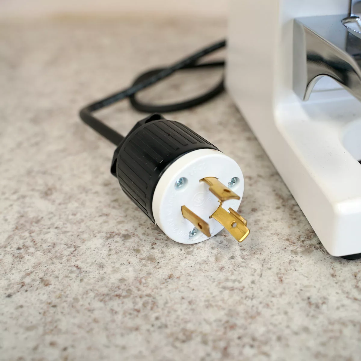 A close-up of the CONVENTION DEMO - Slingshot Kilo Precision Automatic Tamper White 220V plug with gold prongs and black cord, resting on a speckled countertop next to a white appliance. © 2025 1st-line.com
