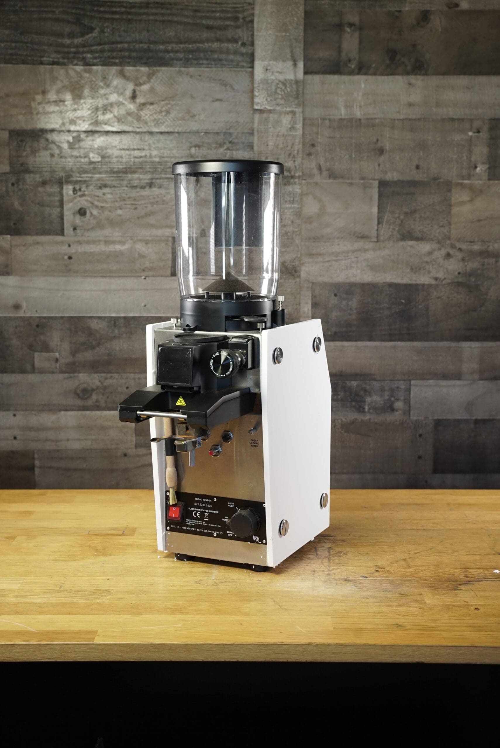 A CONVENTION DEMO - Slingshot Dosis S75 Espresso Coffee Grinder White 220V with a transparent hopper sits on a wooden countertop, featuring control knobs and a black front panel against a wood-paneled wall. © 2025 1st-line.com