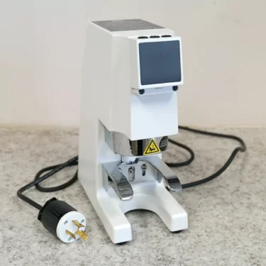 A CONVENTION DEMO - Slingshot Kilo Precision Automatic Tamper White 220V with a display screen, metal parts, and an Australian plug sits on a gray countertop against a light wall. © 2025 1st-line.com