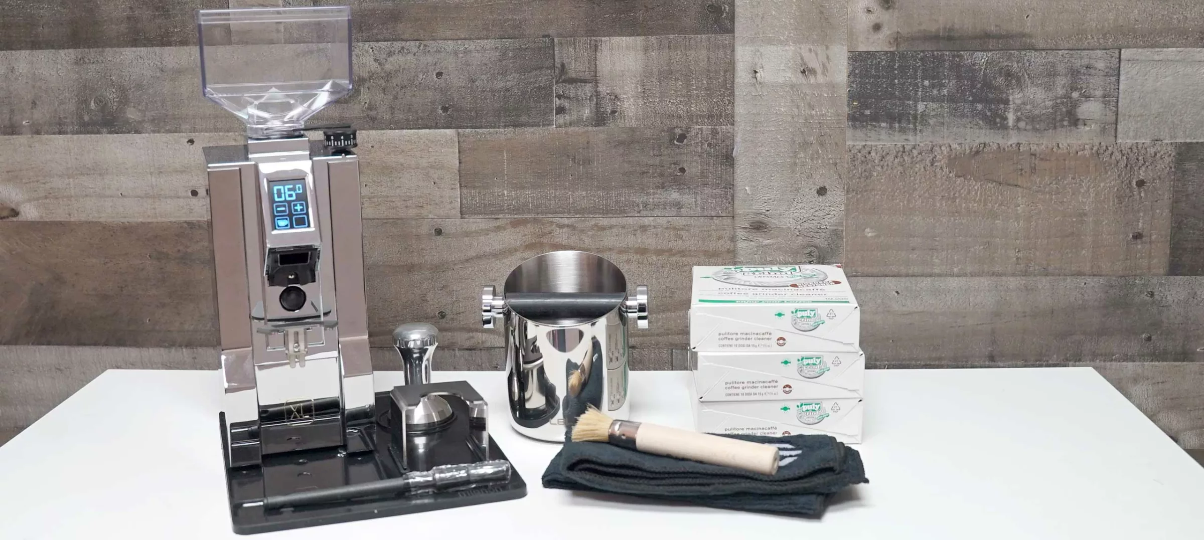 A MignonXL coffee grinder with a digital display sits on a white counter next to a stainless steel knock box, three stacked boxes, a brush, a tamper, and a folded black cloth, all against a wood-paneled wall. © 2025 1st-line.com