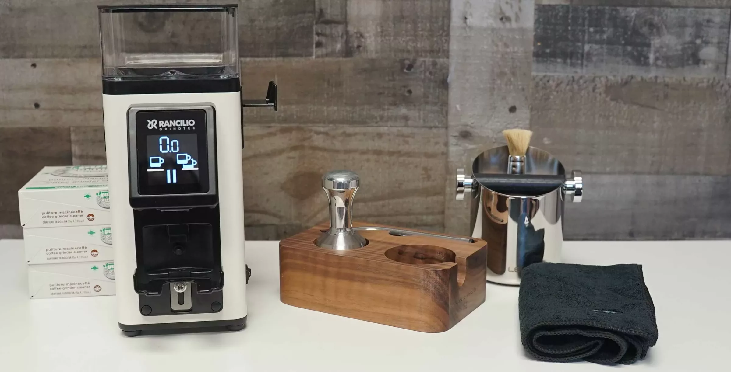 A Stile coffee grinder with a digital display sits on a counter next to a wooden espresso tool holder with a tamper, a metal knock box with a brush, and a folded dark towel. The background features wood paneling. © 2025 1st-line.com