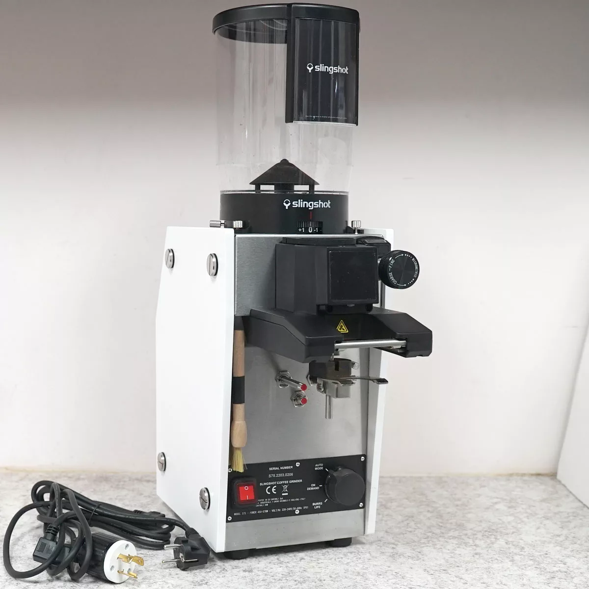A white Slingshot Dosis S75 Espresso Coffee Grinder (CONVENTION DEMO, 220V) with a transparent bean hopper sits on the countertop, its power cord and plug coiled neatly beside it. © 2025 1st-line.com