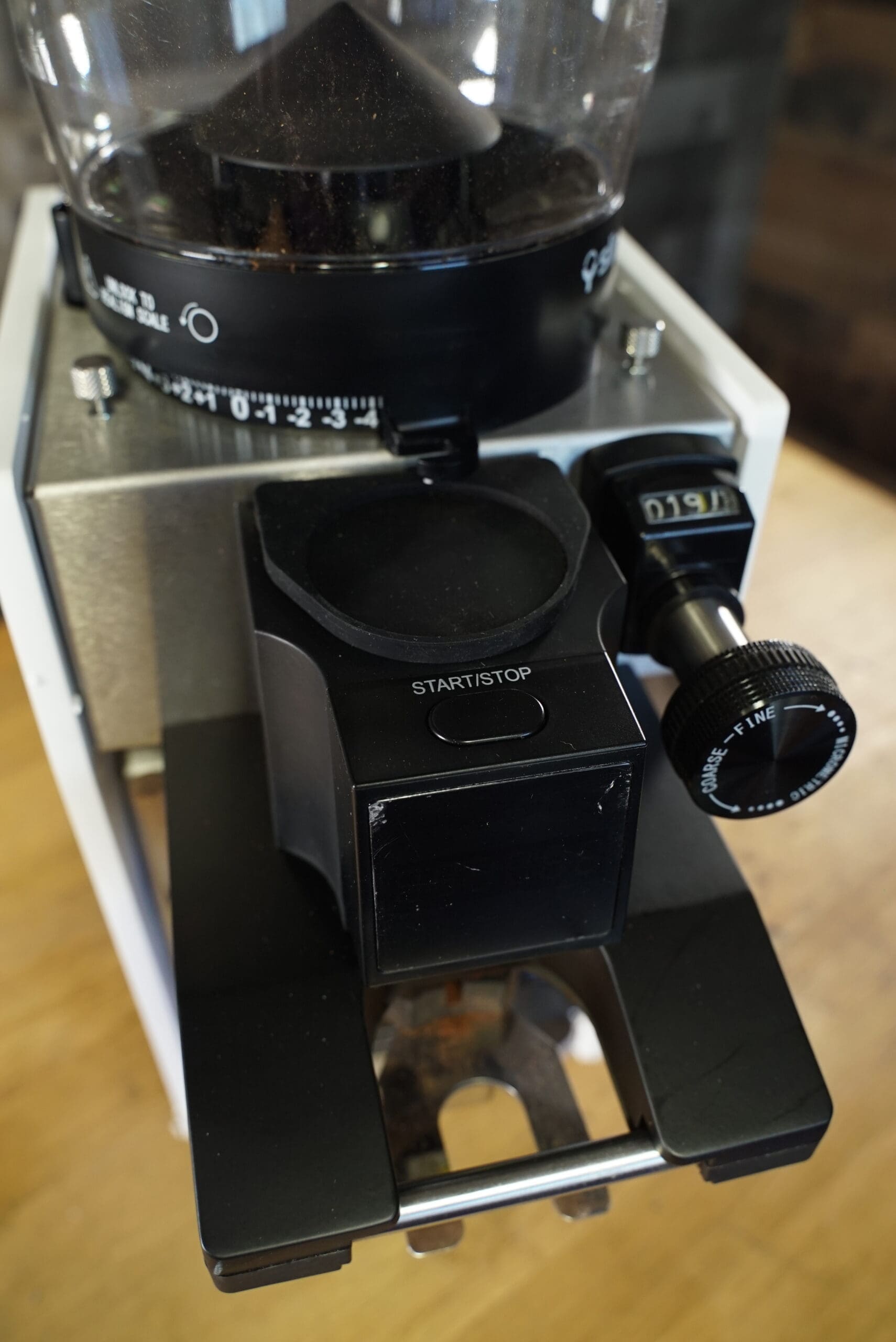 Close-up of a CONVENTION DEMO - Slingshot Dosis S75 Espresso Coffee Grinder White 220V, featuring a black start/stop button, adjustable numbered dial, and transparent hopper, sitting on a wooden surface. © 2025 1st-line.com