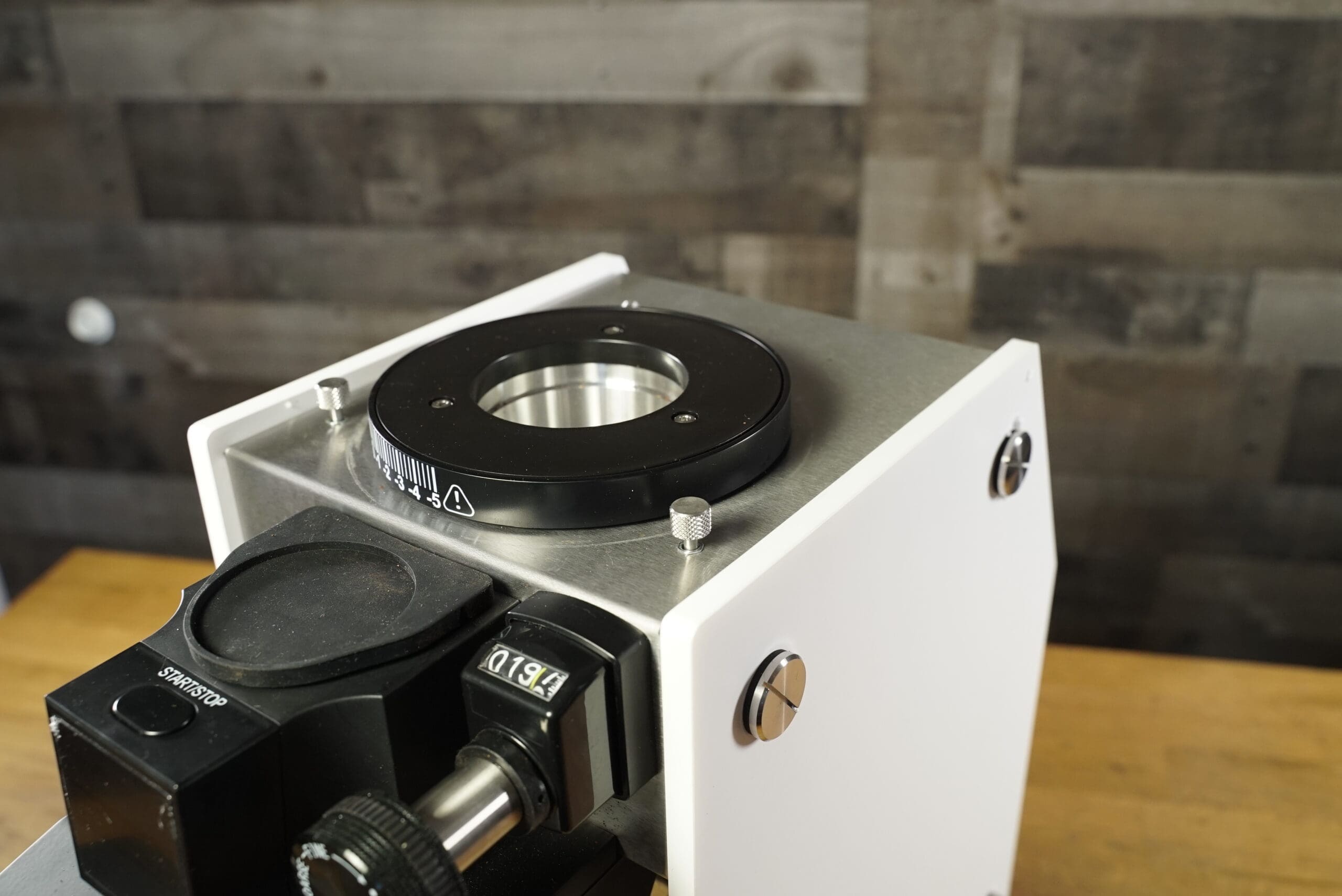 Close-up of the CONVENTION DEMO - Slingshot Dosis S75 Espresso Coffee Grinder White 220V on a wooden surface with dial and adjustment knob, set against wooden wall panels. © 2025 1st-line.com