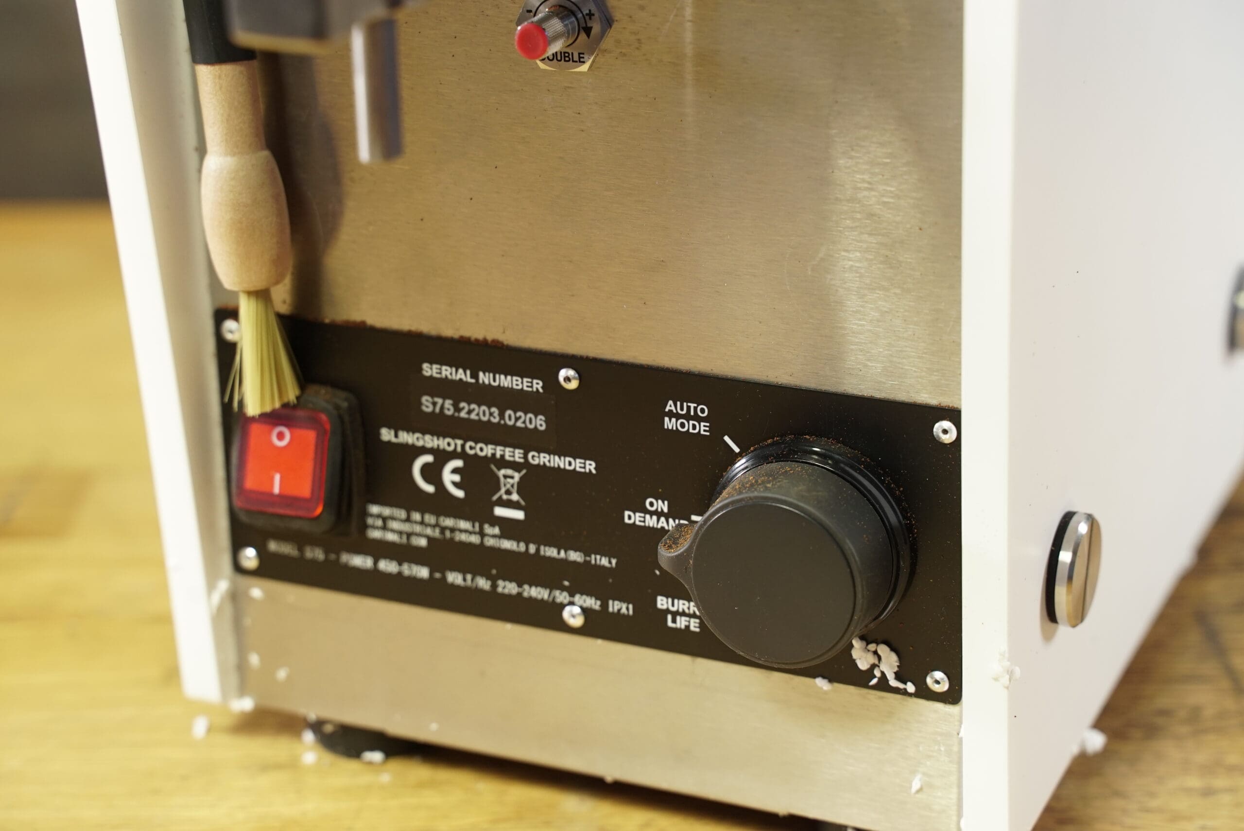 Close-up of the CONVENTION DEMO - Slingshot Dosis S75 Espresso Coffee Grinder White 220V’s control panel, with a red power switch, 220V label, brush on a hook, black adjustment knob, and scattered coffee grounds. © 2025 1st-line.com