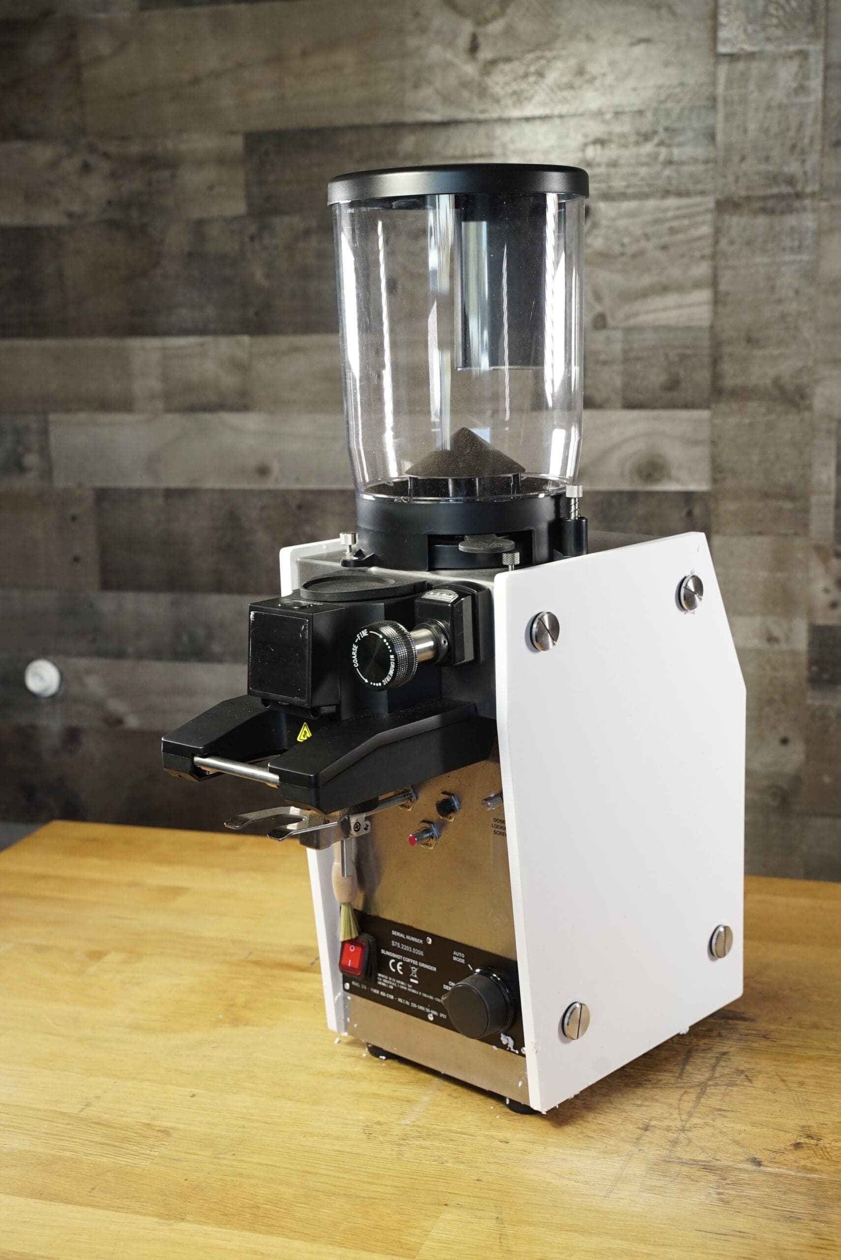 The CONVENTION DEMO - Slingshot Dosis S75 Espresso Coffee Grinder White 220V, featuring a transparent bean hopper, sits on a wooden countertop against a rustic wall, blending seamlessly into any professional café setting. © 2025 1st-line.com
