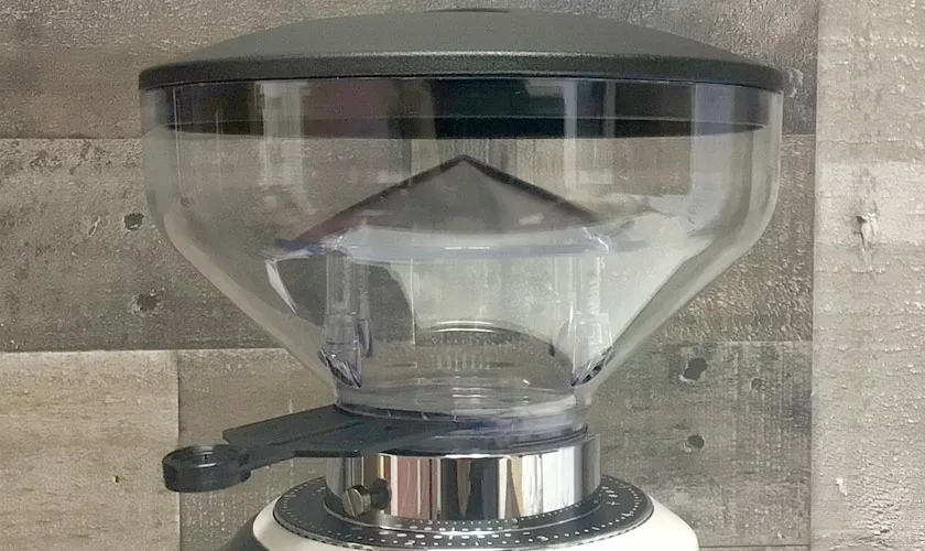 A close-up of a clear plastic hopper with a black lid on the white Quamar Nemo QE Espresso Coffee Grinder, set against a wood-textured background—ideal for upgrading your espresso coffee setup. © 2025 1st-line.com