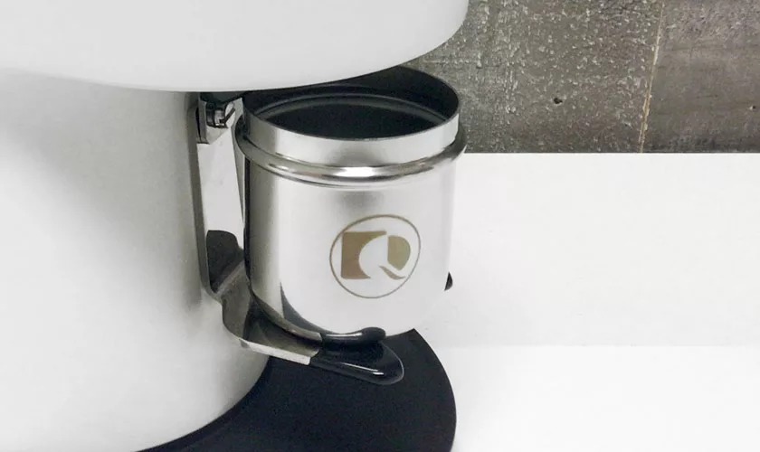 A close-up of a metallic container with a logo, placed beneath the Quamar Nemo QE Espresso Coffee Grinder on a white surface, with a textured gray wall in the background. © 2025 1st-line.com