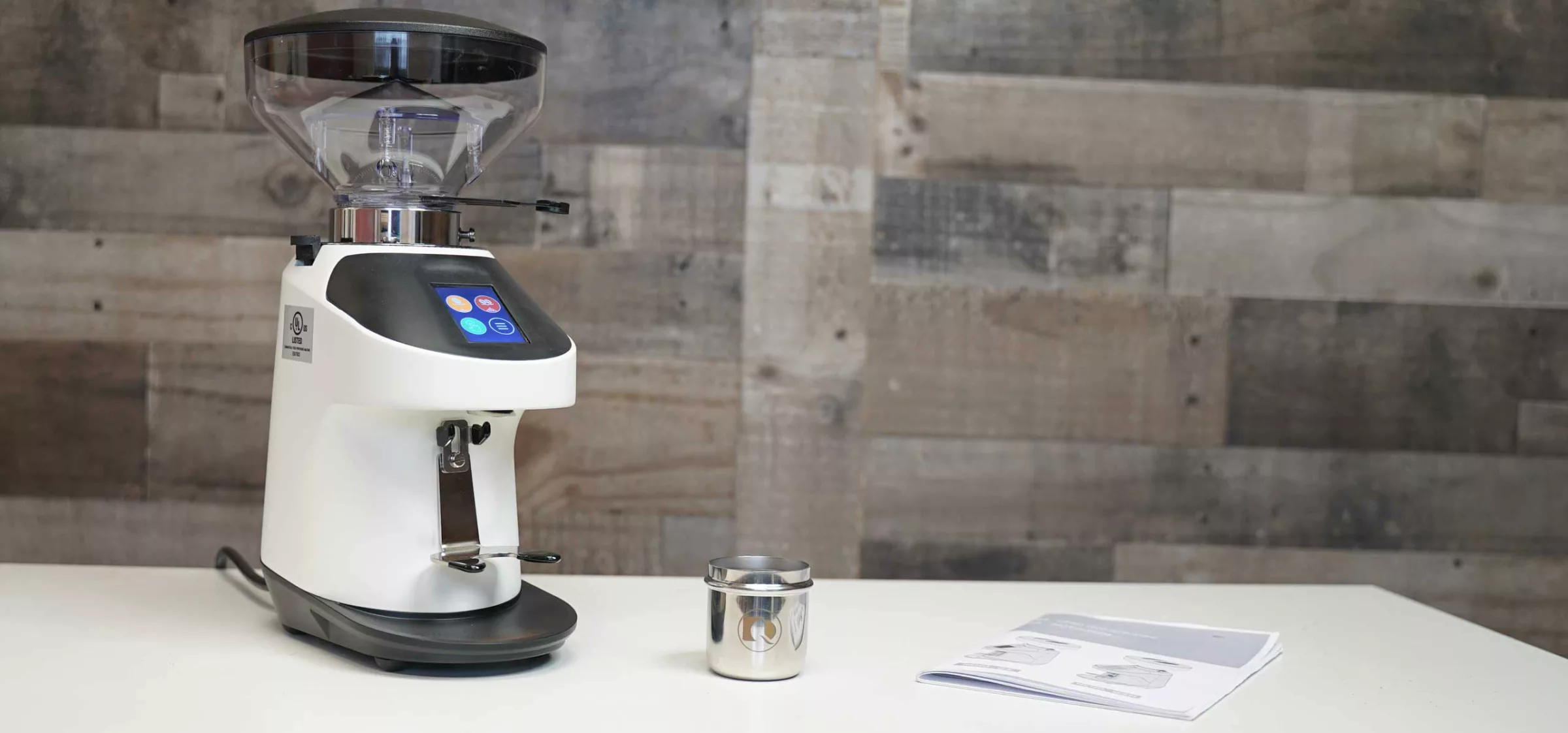A Quamar Nemo QE Espresso Coffee Grinder sits on a white table beside a small metal container and an open manual, with a wooden plank wall in the background. © 2025 1st-line.com