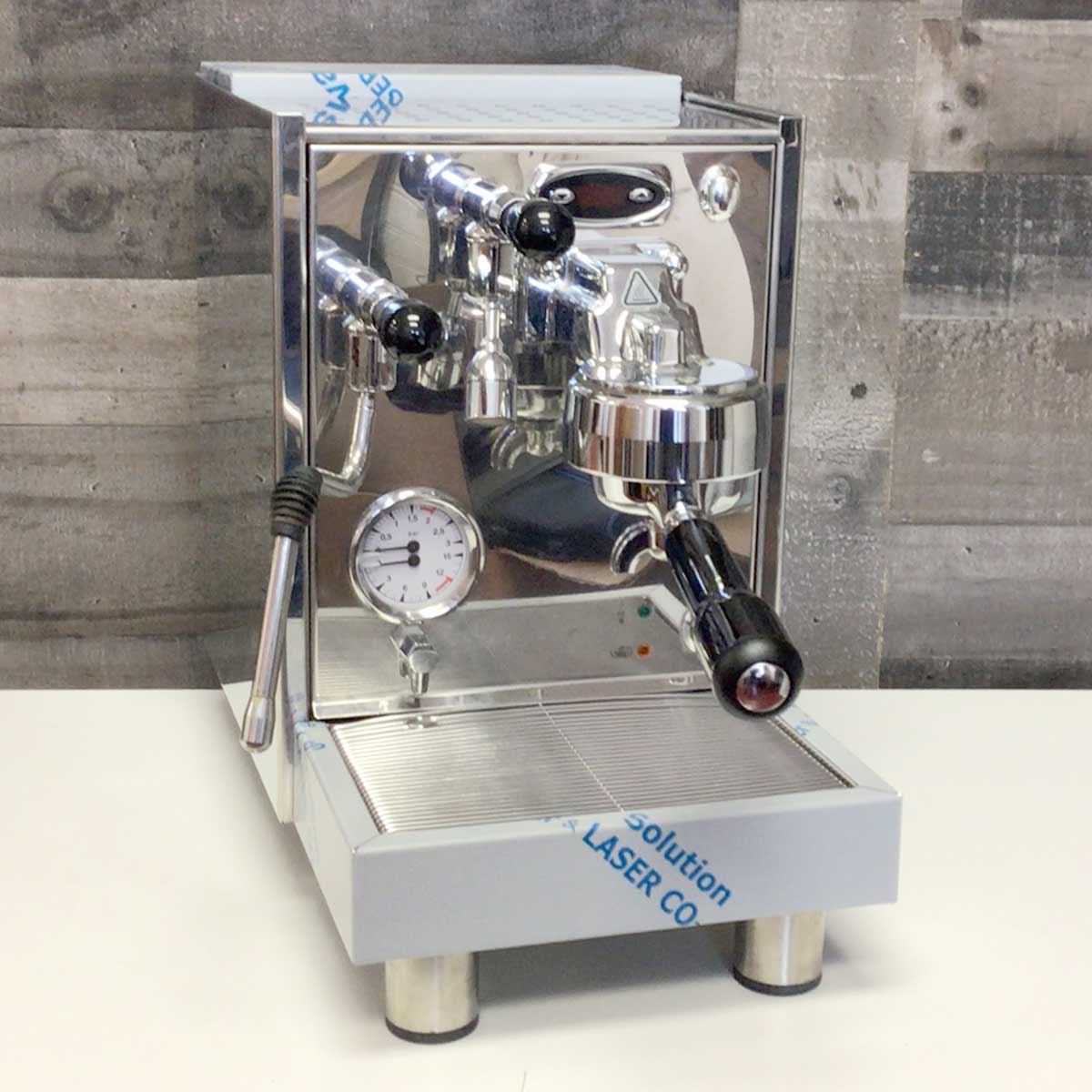 A shiny SIDEWALK SALE - Bezzera BZ07 V1 PM PID Espresso Machine (demo, E112) with pressure gauge, steam wand, and portafilter sits on a white surface against a gray wooden wall background. © 2025 1st-line.com