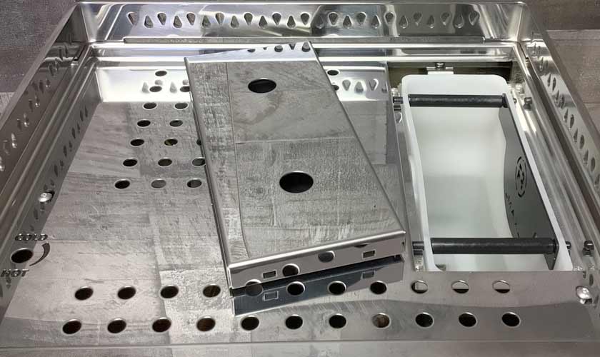 A stainless steel medical sterilization tray with perforated holes lies open, containing rectangular metal containers and a white plastic divider—crafted with precision similar to that of the Rocket Epica Espresso Machine. © 2025 1st-line.com