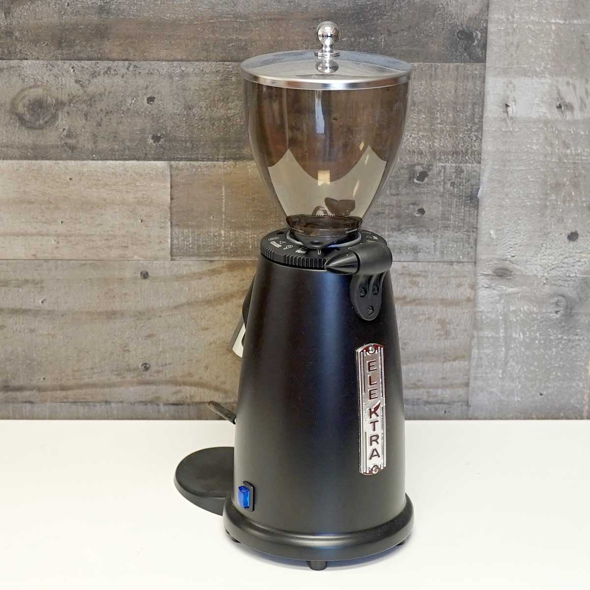 An Elektra MSD Black Espresso Coffee Grinder (demo) (C204) with a clear bean hopper is placed on a white surface in front of a wood-patterned wall. © 2025 1st-line.com