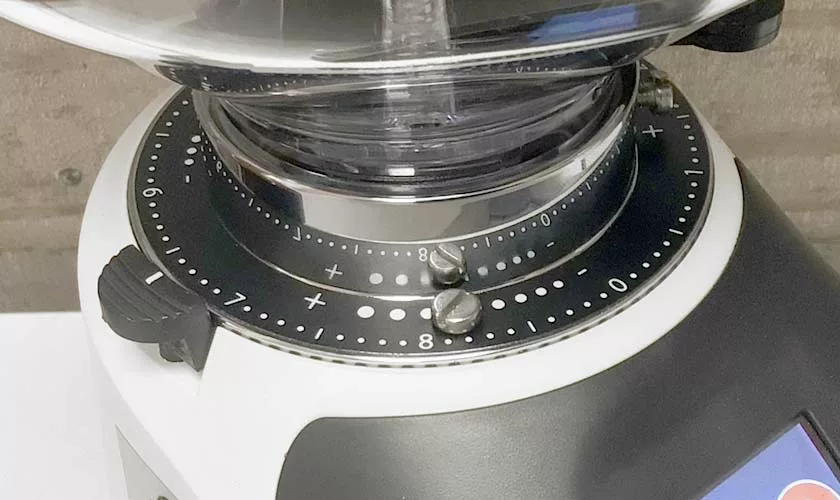 Close-up of the Quamar Nemo QE Espresso Coffee Grinder highlights its grind size adjustment dial with numbered and dotted markings, a clear hopper on top, and part of a digital display visible on the side. © 2025 1st-line.com