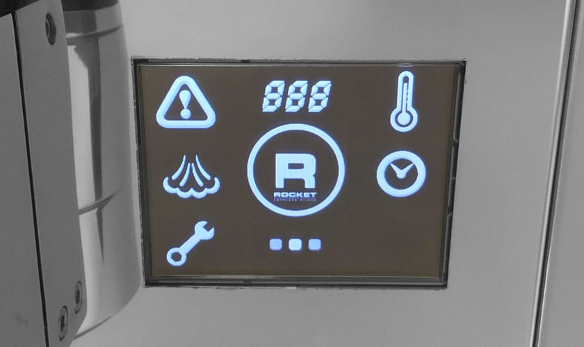 The Rocket Epica Espresso Machine features a digital control panel with backlit white icons—warning sign, steam, wrench, thermometer, clock, and a bold “R” Rocket logo—all shown on a dark background. © 2025 1st-line.com