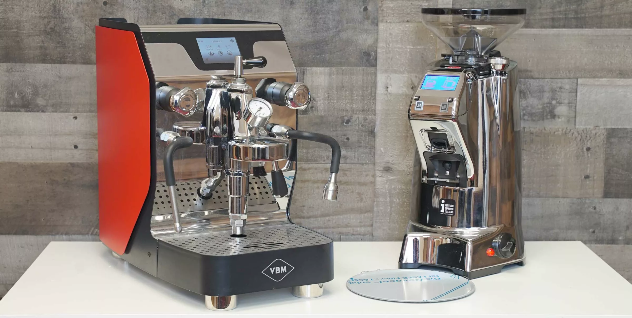 The *SOLD* VBM Dual Boiler / Eureka Zenith 65E Duo, featuring a red-panel espresso machine and chrome grinder, sits on a white counter against a wooden wall. © 2025 1st-line.com
