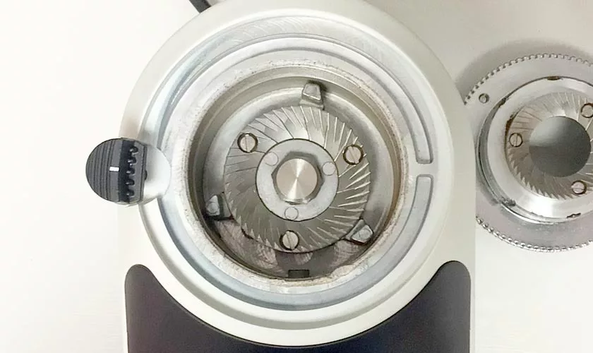 Top-down view of a Quamar Nemo QE Espresso Coffee Grinder with its burr exposed, revealing sharp metal blades and a black adjustment knob to the left. A detached burr is placed on the right side on a white surface. © 2025 1st-line.com