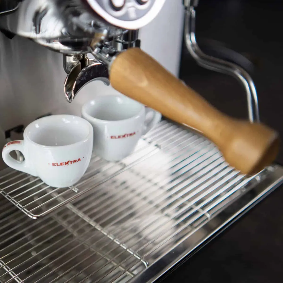 Close-up of the Elektra Evok 2 Group Commercial Espresso Machine with a wooden-handled portafilter. Two white ceramic cups labeled ELEKTRA rest on the drip tray, positioned beneath the spout, ready for coffee. 2025 1st-line.com