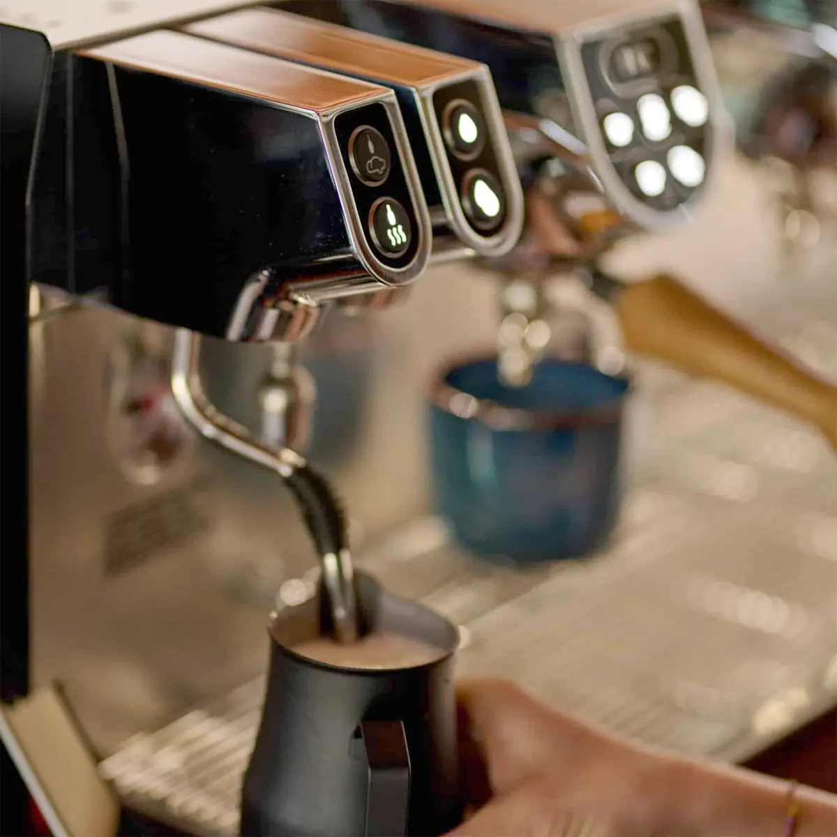 Close-up of a person steaming milk with a stainless steel pitcher using the Elektra Evok 2 Group Commercial Espresso Machine, with buttons and a cup visible in the background. 2025 1st-line.com