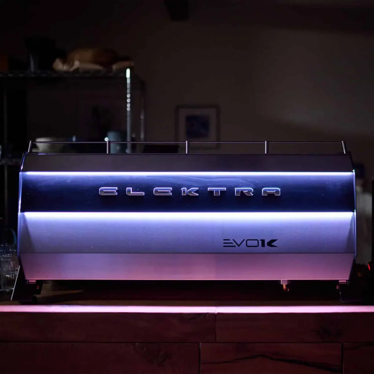 The Elektra Evok 2 Group Commercial Espresso Machine features blue and purple LED lighting, illuminating its sleek design as it sits on a wooden counter in a dimly lit room. 2025 1st-line.com