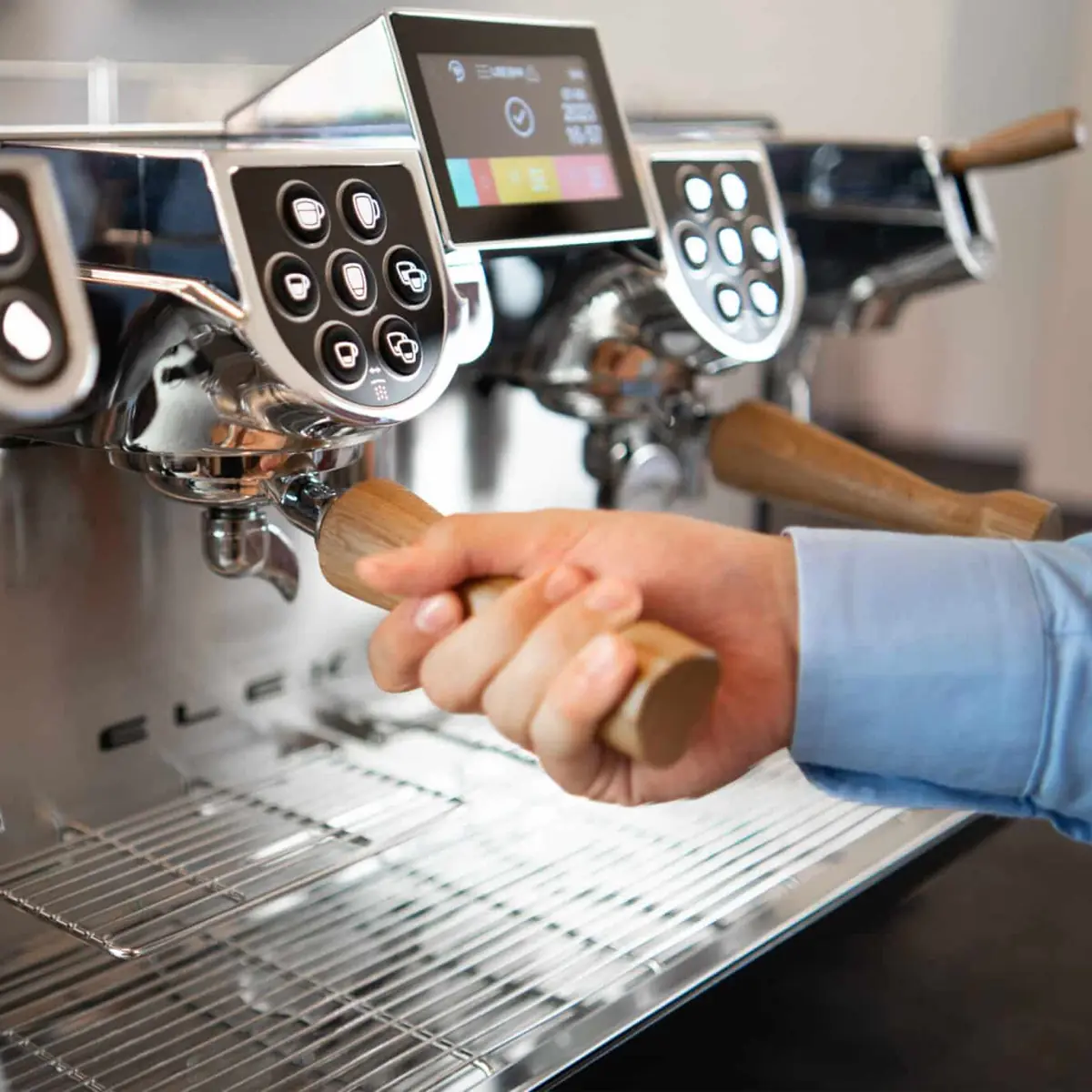 A person in a blue shirt prepares coffee using the Elektra Evok 2 Group Commercial Espresso Machine, gripping its wooden portafilter handle and utilizing its digital displays and multiple control buttons. 2025 1st-line.com