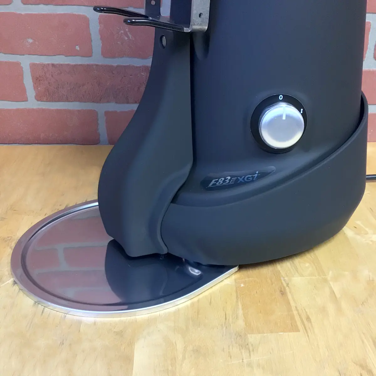 A close-up of a black Fiorenzato F83 E XGi Espresso Coffee Grinder with a silver dial and metal tray, placed on a wooden surface in front of a red brick wall. © 2025 1st-line.com