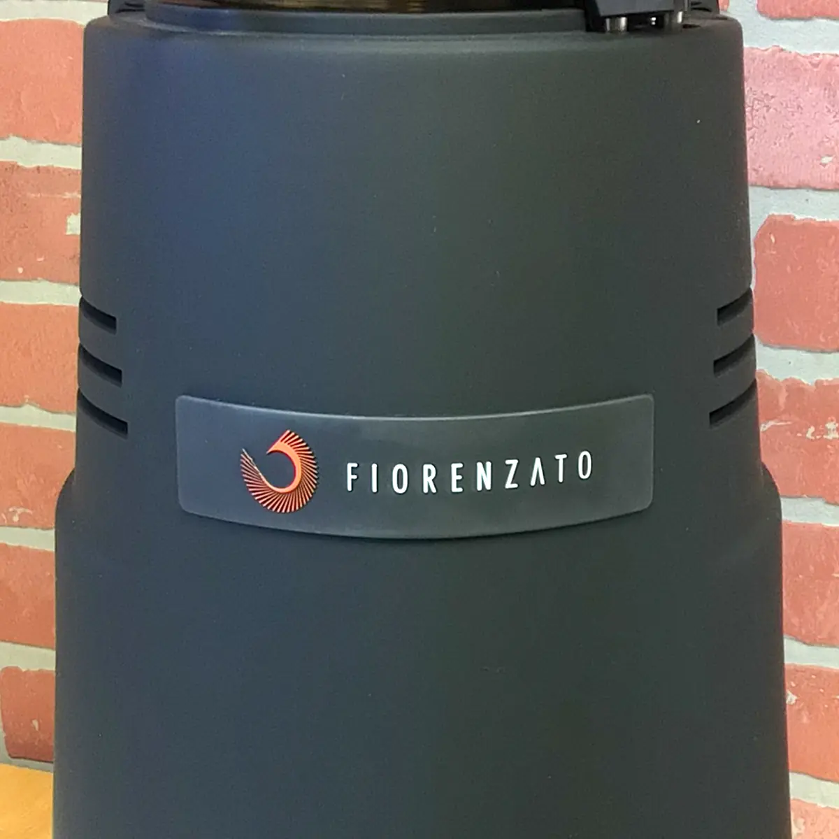 A close-up of a matte black Fiorenzato F83 E XGi Espresso Coffee Grinder with a stylized red logo and white text on a gray label, set against a pinkish brick wall background. © 2025 1st-line.com