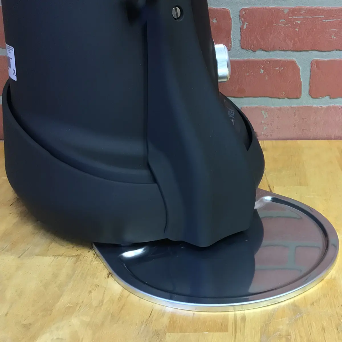 A black Fiorenzato F83 E XGi Espresso Coffee Grinder sits on a wooden countertop against a brick wall. A shiny, round metal plate at its base reflects the surroundings. © 2025 1st-line.com