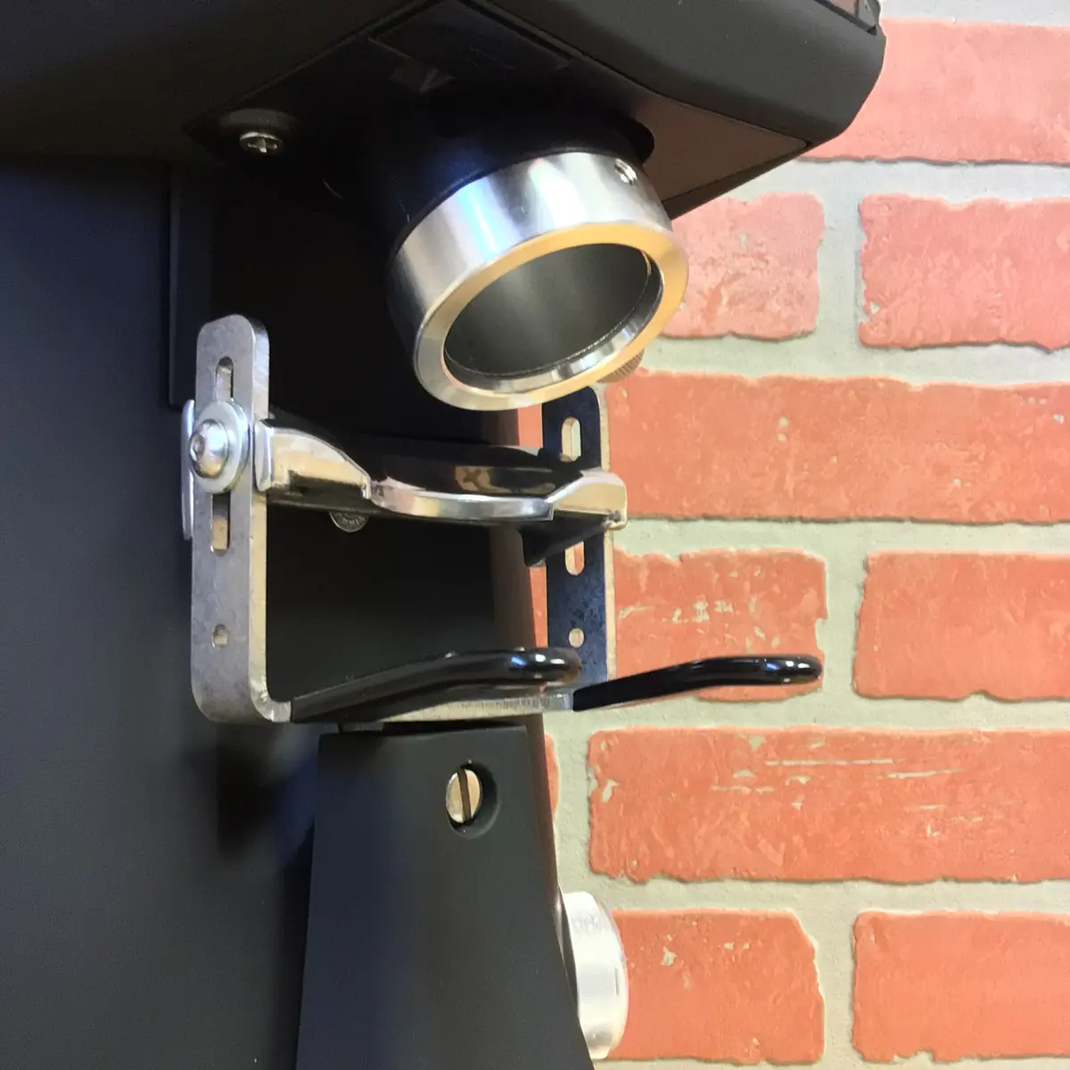 A close-up of a portafilter holder attached to a Fiorenzato F83 E XGi Espresso Coffee Grinder, set against red brick-patterned wallpaper, with the portafilter clamp and grinder spout in view. © 2025 1st-line.com