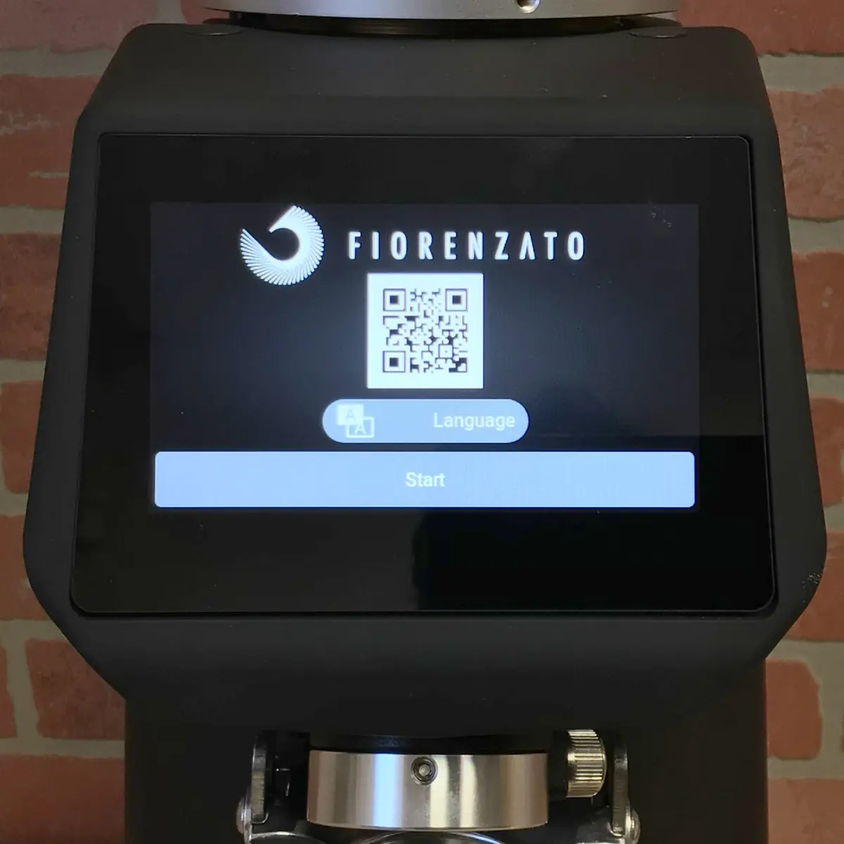 The Fiorenzato F83 E XGi Espresso Coffee Grinder’s touchscreen displays the Fiorenzato logo, a QR code, language selection options, and a Start button on a brick wall background. © 2025 1st-line.com