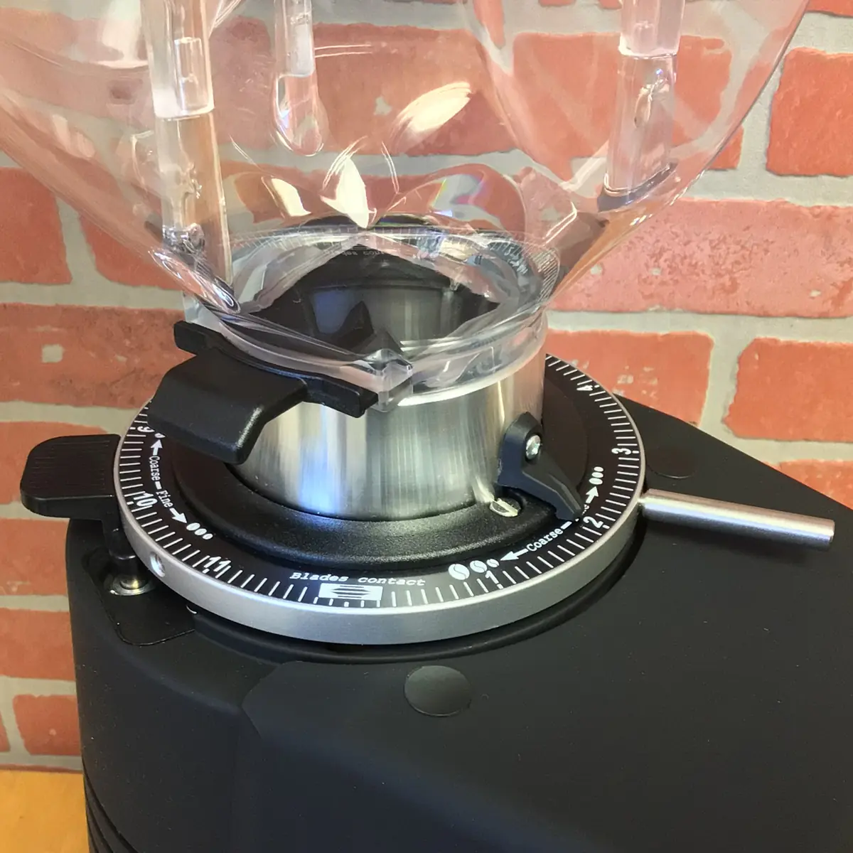Close-up of a Fiorenzato F83 E XGi Espresso Coffee Grinder adjustment dial with numbers and lever, set against a red brick wall. The transparent hopper and black body of the grinder are visible. © 2025 1st-line.com