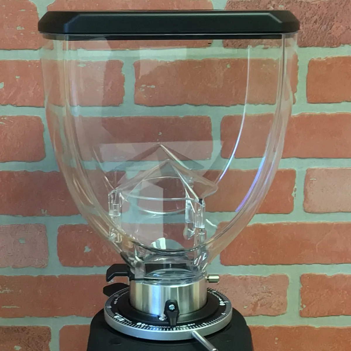 A clear, empty plastic hopper with a black lid sits atop a Fiorenzato F83 E XGi Espresso Coffee Grinder against a red brick wall background. © 2025 1st-line.com