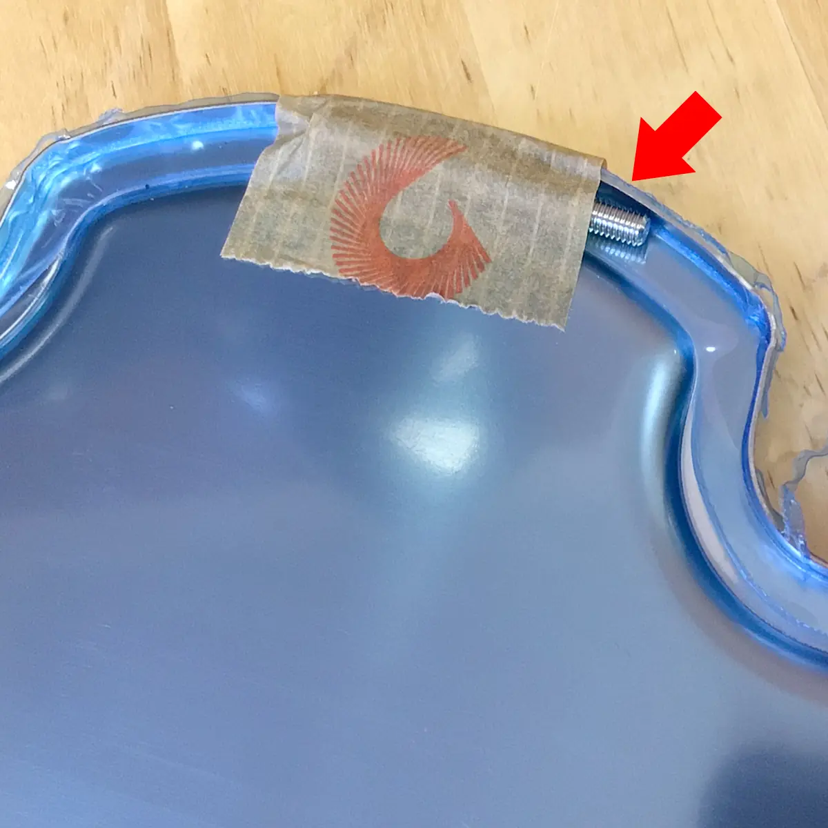 A close-up of a blue Fiorenzato F83 E XGi Espresso Coffee Grinder part with a metal screw partly inserted at the edge, held by tape. A red arrow points to the screw. The part rests on a wooden background. © 2025 1st-line.com